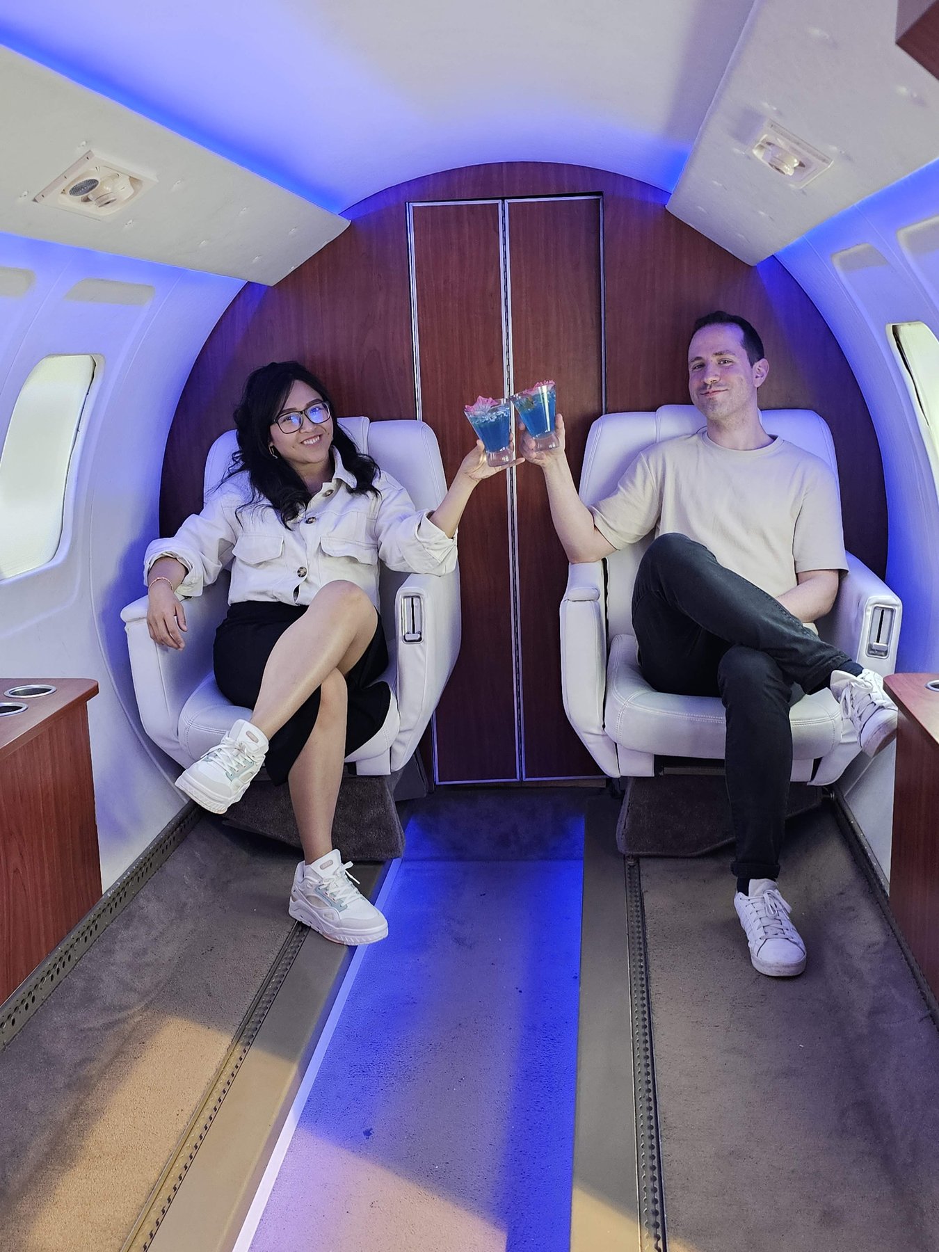 Chern and Javier toasting with blue cocktails inside a softly glowing airplane cabin